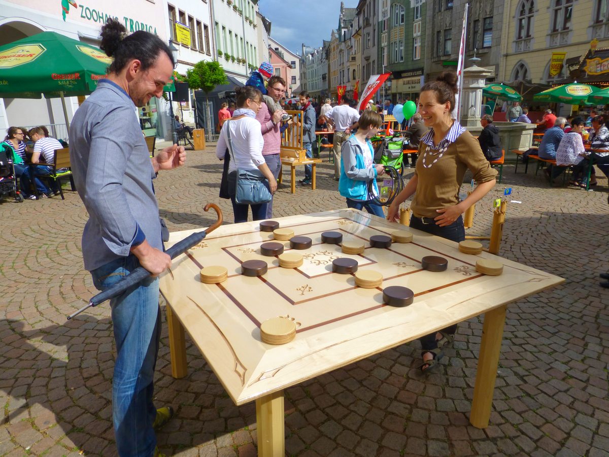 Zwei Gäste spielen an einem XXL-Holzspiel auf einem belebten Marktplatz